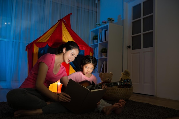 jovem mãe linda segurando o livro de partituras acompanhar linda menina bonita tocando ukulele de guitarra pequena no quarto de cama com barraca de crianças à noite.