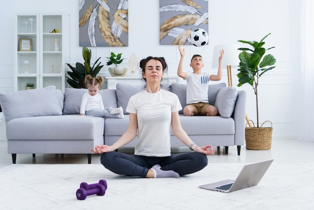 Jovem mãe meditando na posição de ioga de lótus, enquanto seus filhos brincando em casa