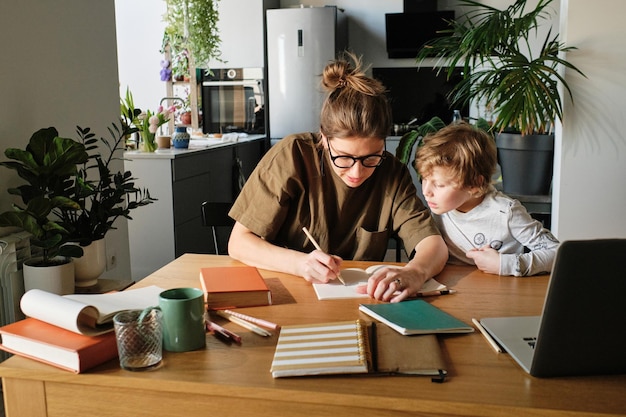 Jovem mãe mostrando como escrever para seu filho enquanto eles fazem lição de casa juntos na mesa na cozinha