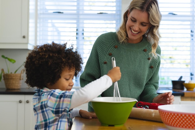 Foto jovem mãe multirracial sorridente olhando para o filho batendo massa em uma tigela na mesa na cozinha