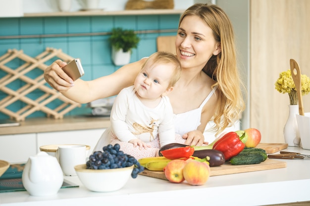 Jovem mãe olhando para a câmera e sorrindo, cozinhando e brincando com sua filha bebê em uma cozinha moderna. usando o telefone, fazendo selfie.