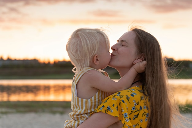 Jovem mãe segurando a filha e a beija suavemente. Retrato por do sol no fundo do rio.