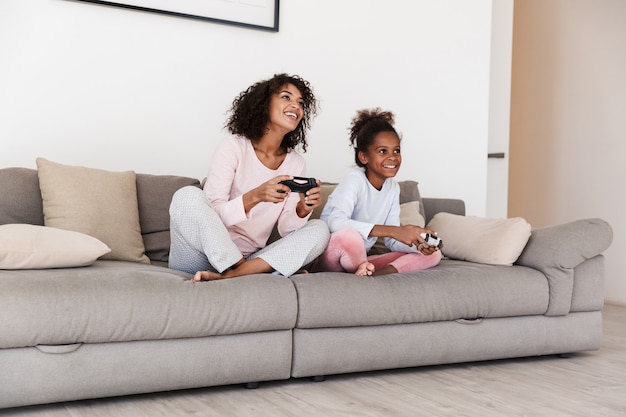 Jovem mãe sorridente e sua filha de pijama relaxando no sofá, jogando videogame