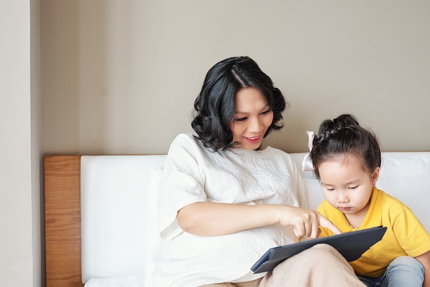 Jovem mãe sorridente mostrando um aplicativo educacional em um computador tablet para sua filha