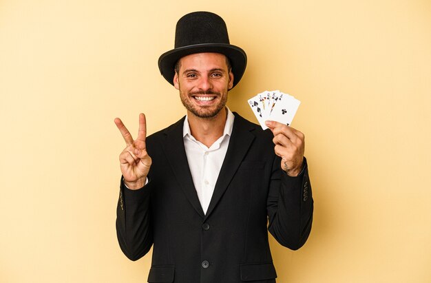 Jovem mago caucasiano segurando cartão mágico isolado em fundo amarelo, mostrando o número dois com os dedos.