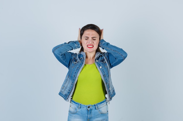 Jovem, mantendo as mãos nas orelhas em roupa jeans e parecendo irritada, vista frontal.