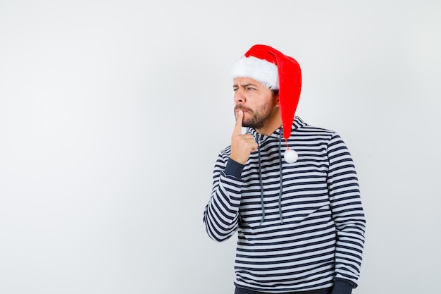 Jovem mantendo o dedo no queixo, olhando para longe com um capuz, chapéu de Papai Noel e parecendo pensativo,