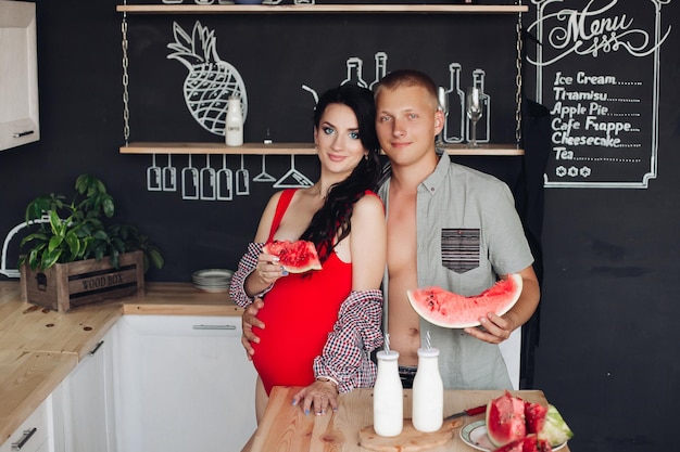 Jovem marido abraçando sua linda esposa grávida de blusa vermelha e camisa Eles segurando melancia fatiada na mesa de madeira na cozinha moderna
