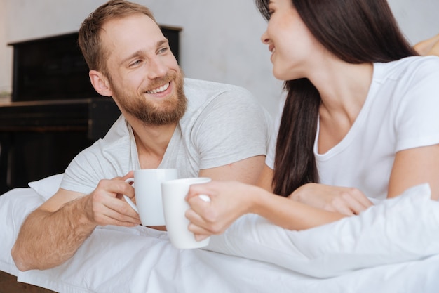 Jovem marido e mulher deitados na cama com uma xícara de café e conversando sobre o travesseiro pela manhã