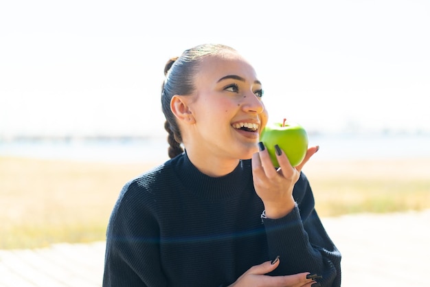 Jovem marroquina ao ar livre segurando uma maçã com expressão feliz