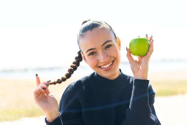 Jovem marroquina ao ar livre segurando uma maçã com expressão feliz