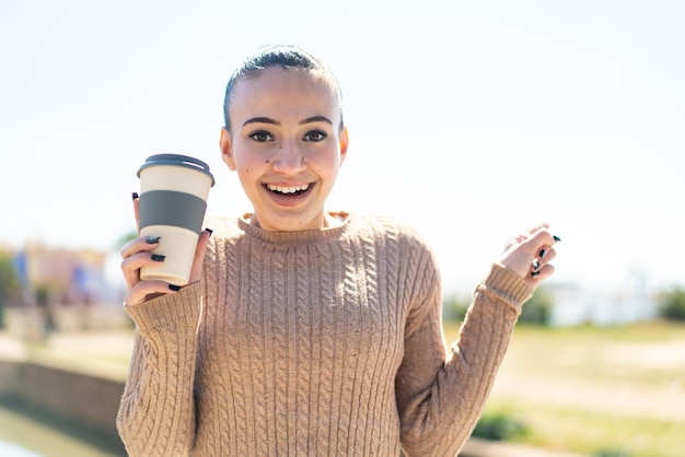 Jovem marroquina segurando um café para viagem ao ar livre surpreso e apontando o dedo para o lado