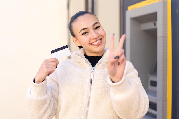 Jovem marroquina segurando um cartão de crédito ao ar livre sorrindo e mostrando sinal de vitória