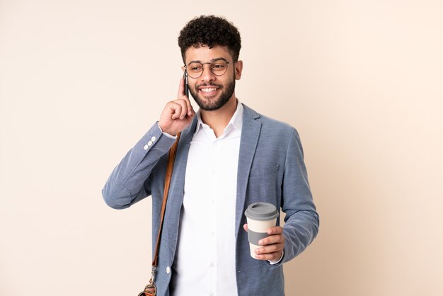 Jovem marroquino, empresário, isolado em um bege, segurando um café para levar e um celular