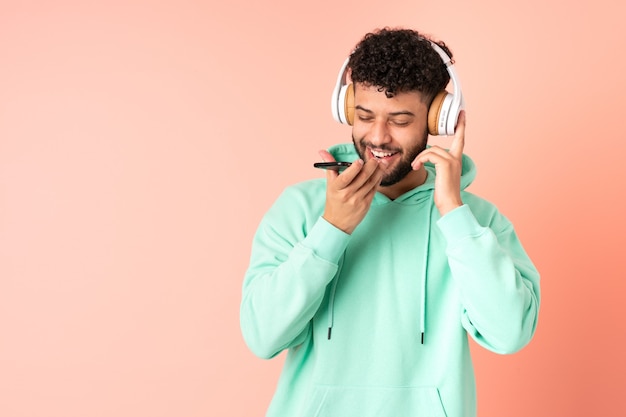 Jovem marroquino isolado em uma parede rosa ouvindo música com um celular e cantando