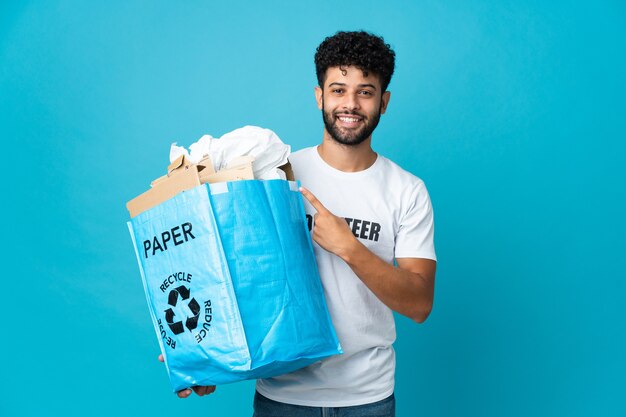 Jovem marroquino segurando uma sacola de reciclagem cheia de papel para reciclar sobre uma parede isolada apontando para o lado para apresentar um produto