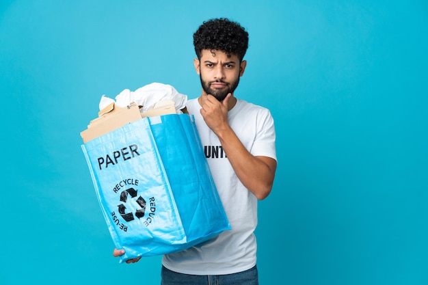 Foto jovem marroquino segurando uma sacola de reciclagem cheia de papel para reciclar sobre uma parede isolada com dúvidas