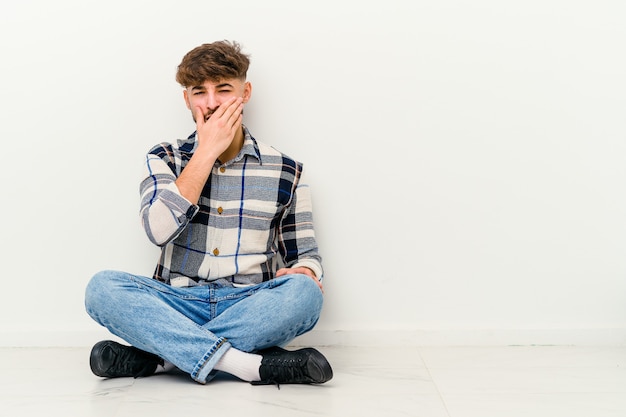 Jovem marroquino sentado no chão, isolado na parede branca, rindo de emoção feliz, despreocupada e natural.