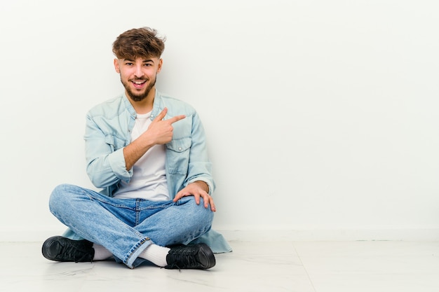 Jovem marroquino sentado no chão, isolado na parede branca, sorrindo e apontando de lado, mostrando algo no espaço em branco.