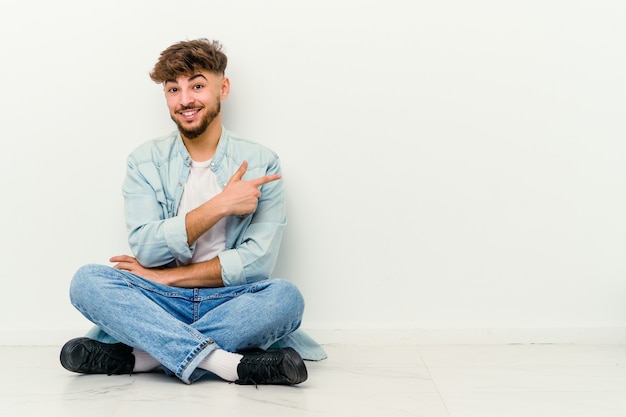 Jovem marroquino sentado no chão isolado no branco, sorrindo alegremente, apontando com o dedo indicador para longe.