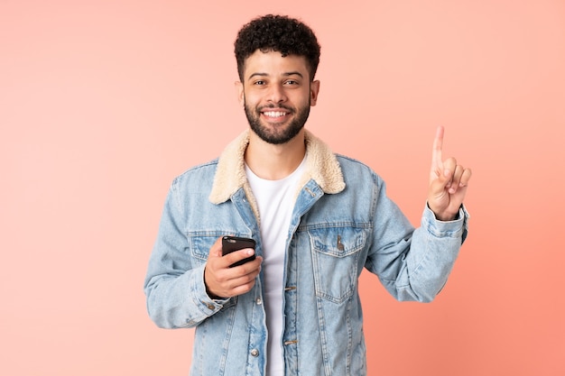 Jovem marroquino usando telefone celular isolado em uma parede rosa apontando para uma ótima ideia