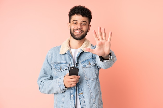 Jovem marroquino usando telefone celular isolado na parede rosa, contando cinco com os dedos