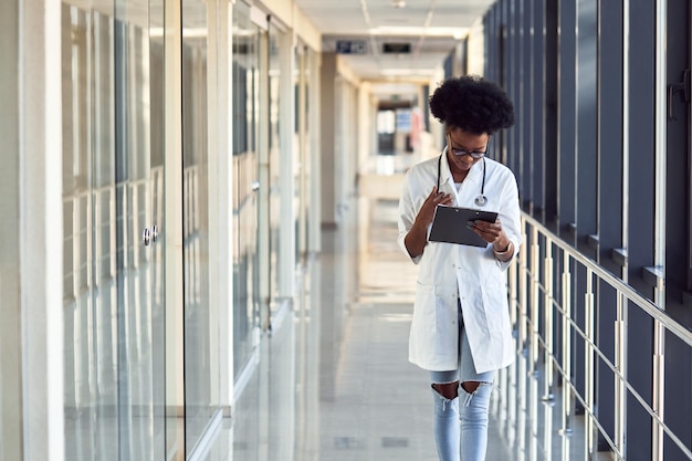 Jovem médica afro-americana de uniforme branco com estetoscópio e bloco de notas em pé no corredor.