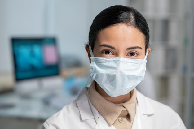 Jovem médica bonita de jaleco branco e máscara protetora no rosto