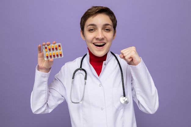 Jovem médica com jaleco branco com estetoscópio em volta do pescoço segurando a bolha com comprimidos feliz e animada com o punho cerrado em pé no roxo