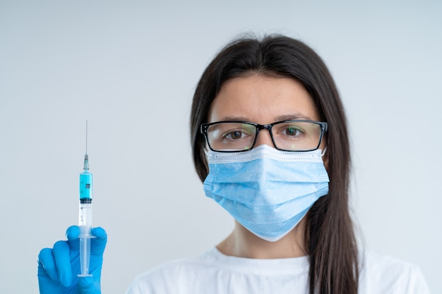 Jovem médica com uma máscara médica e luvas segurando uma seringa com uma vacina contra o coronavírus