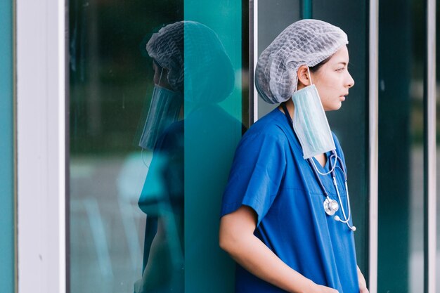 Jovem médica de fato de proteção, uniforme e máscara de rosto, exausta e preocupada.