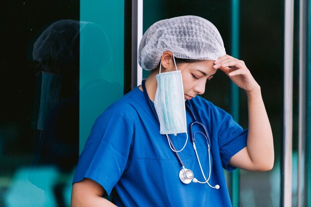 Jovem médica de fato de proteção, uniforme e máscara de rosto, exausta e preocupada.