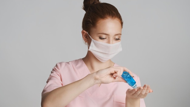 Jovem médica em máscara médica aplicando anti-séptico sobre fundo branco