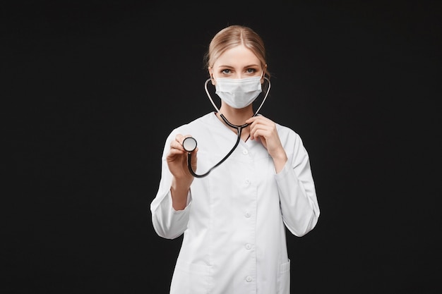 Jovem médica em uniforme médico e máscara protetora usando um estetoscópio