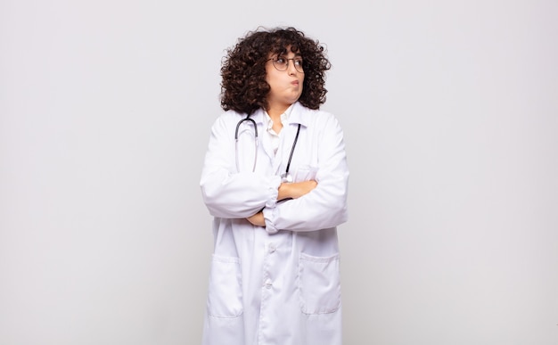 Foto jovem médica encolhendo os ombros, sentindo-se confusa e insegura, duvidando com os braços cruzados e olhar perplexo