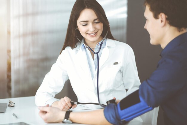 Jovem médica está verificando a pressão arterial de seu paciente, enquanto está sentado juntos na clínica. Conceitos de medicina e saúde.