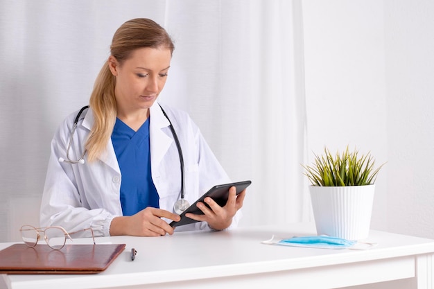 Jovem médica falando com paciente online com tablet digital. Consulta, videochamada, distância. Medicina, saúde durante o Covid-19. Médico com jaleco branco, estetoscópio, pronto para ajudar.