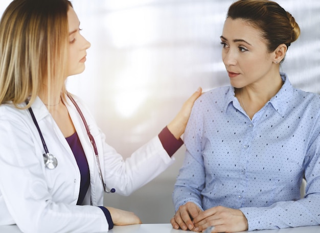 Jovem médica inteligente e um paciente estão discutindo os sintomas de um resfriado do paciente, enquanto estão sentados juntos à mesa no armário ensolarado em uma clínica. A médica está usando uma prancheta e