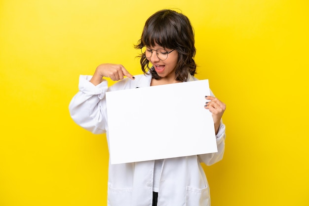 Jovem médica latina isolada em fundo amarelo segurando um cartaz vazio com expressão feliz e apontando-o