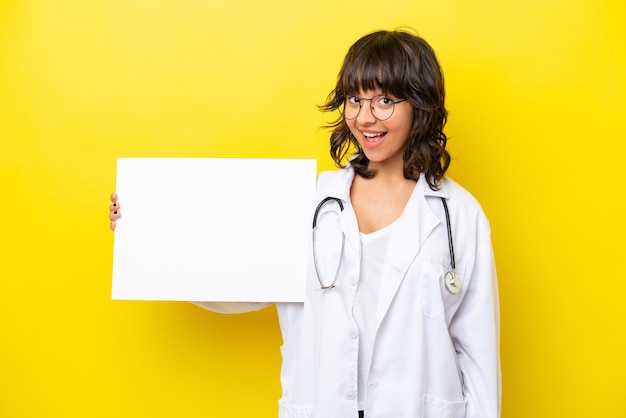 Jovem médica latina isolada em fundo amarelo segurando um cartaz vazio com expressão feliz