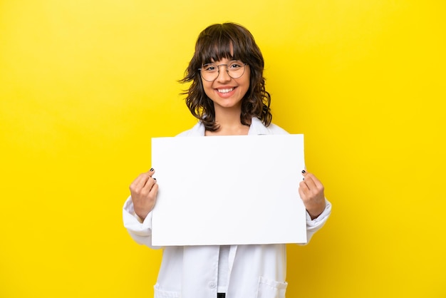 Jovem médica latina isolada em fundo amarelo segurando um cartaz vazio com expressão feliz