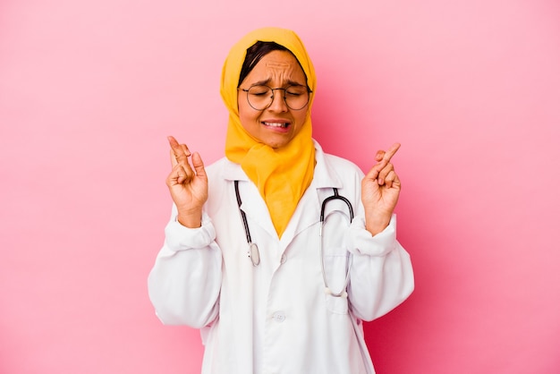 Jovem médica muçulmana isolada em uma parede rosa cruzando os dedos para ter sorte