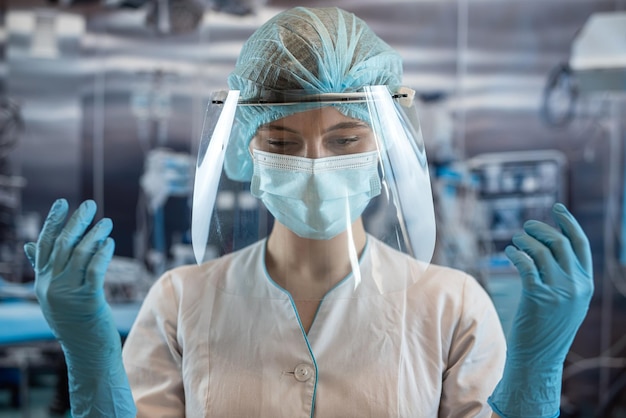 Jovem médica ou enfermeira cirurgiã assistente calçar luvas de proteção azuis em pé na sala de operação Medicina e conceito de saúde