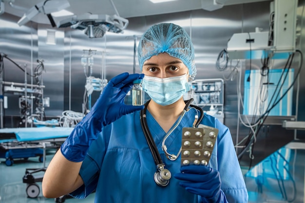 Jovem médica prety vestindo uniforme de casaco azul segurando estetoscópio e pílulas na mão em pé na sala de cirurgia no hospital