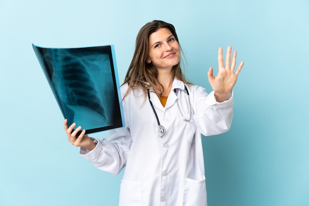 Foto jovem médica segurando uma radiografia sobre uma parede isolada, contando cinco com os dedos