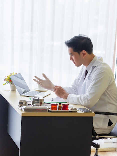 Jovem médico asiático usando luvas de borracha branca em uma mesa que tem um computador