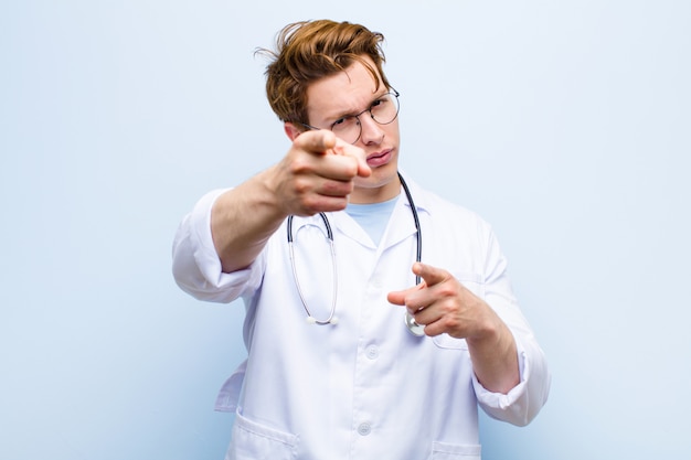 Jovem médico chefe vermelho apontando para a frente com os dois dedos e expressão de raiva, dizendo para fazer seu dever contra a parede azul