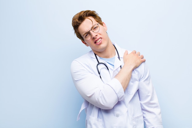 Jovem médico de cabeça vermelha, sentindo-se ansioso, doente, doente e infeliz, sofrendo uma dor de estômago dolorosa ou gripe na parede azul