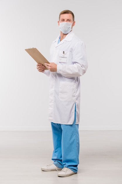 Jovem médico de jaleco e máscara protetora segurando uma prancheta com um documento médico isolado