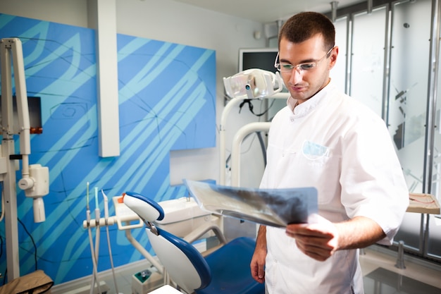 Jovem médico dentista com uniforme branco e óculos em pé olhando para a foto do dente